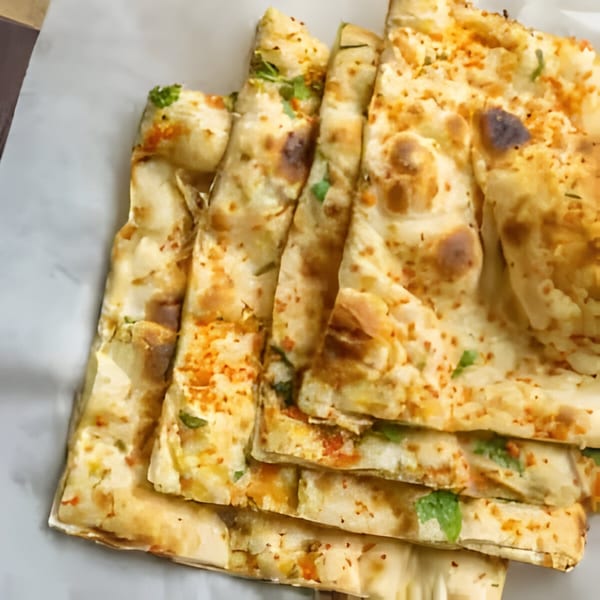 Close-up of golden-brown Indian naan bread slices, garnished with herbs and spices, on white paper.