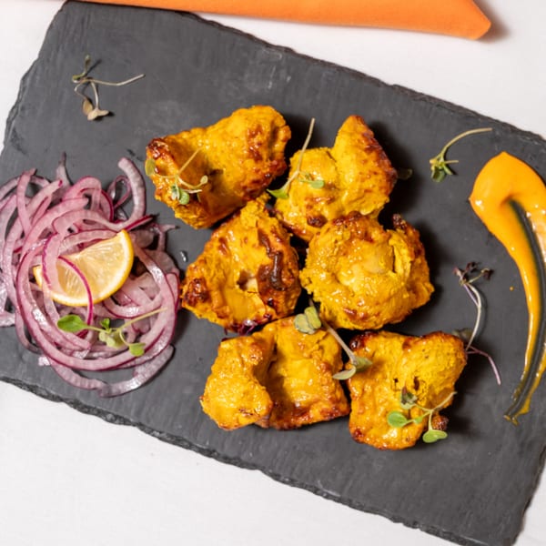 Golden Indian chicken tikka pieces with red onions, lemon, and orange sauce on a dark slate.