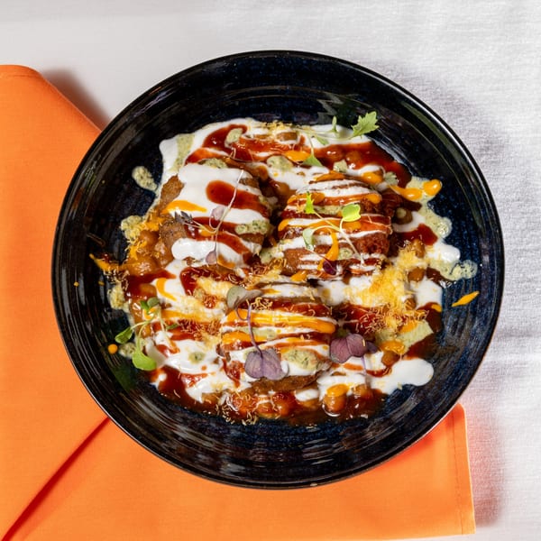 Indian chaat dish with fried patties, yogurt, chutneys, and spices in a dark bowl.